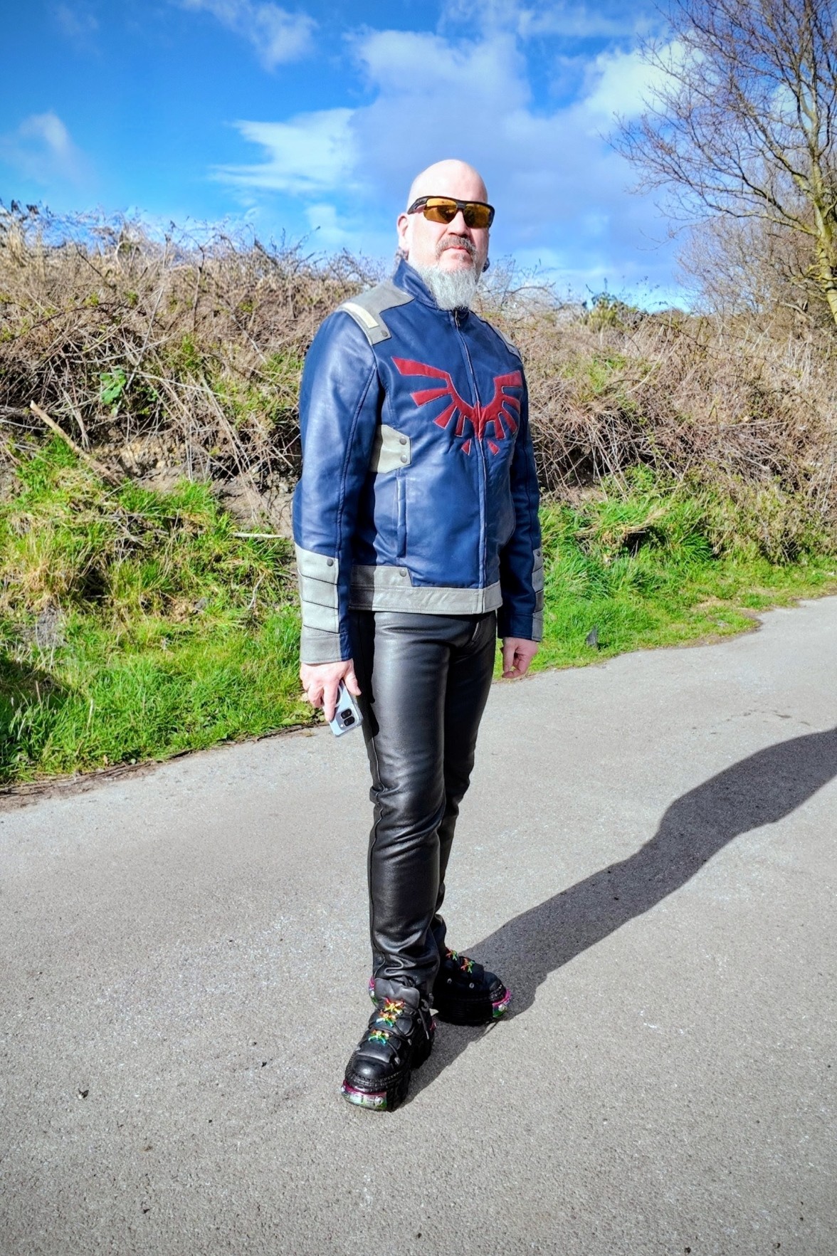I'm standing in a lane showing off my new leather jacket. This is the front view showing a Hyulan crest logo in gold. I'm wearing black leather jeans and new rock boots and I've got a bald head and white beard and I'm wearing Oakley shades. The sky is blue behind but there's a lot of rain bearing clouds building up.