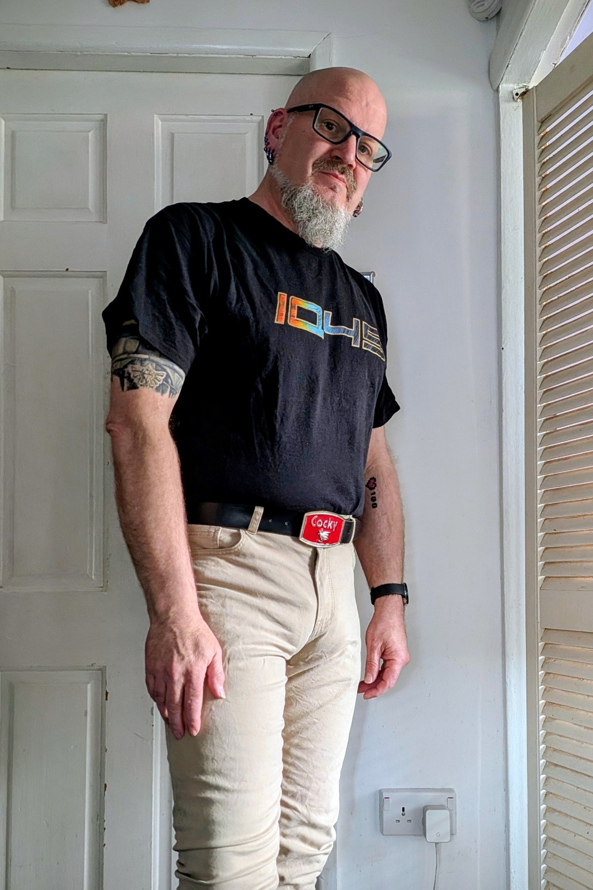 I'm a 52 year old man with a bald head, glasses, white beard and lots of piercings. I'm wearing a t-shirt with IQ45 written on it and I'm in slim fit beige jeans. My belt buckle says "Cocky" on it.
